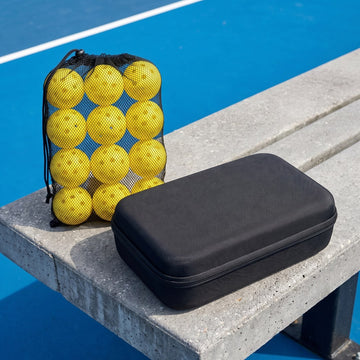 Mesh bag of twelve yellow pickleballs beside a black hardshell paddle case on a courtside bench